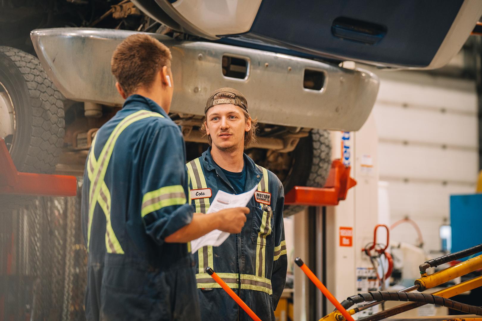 Motion Machinery technicians reviewing an inspection sheet under a truck on the hoist